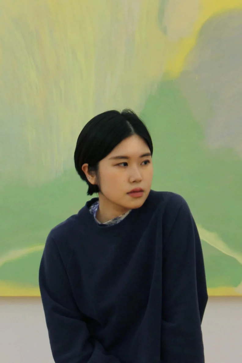Young woman with short black hair and navy sweater sits before a soft green and yellow abstract painting, looking rightward pensively.