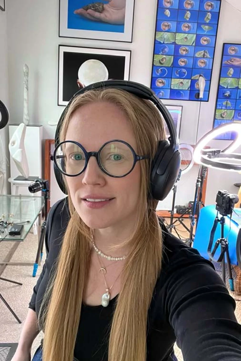 Person with long blonde hair wearing round glasses, black over-ear headphones and pearl necklace, smiling in a studio with ring lights.