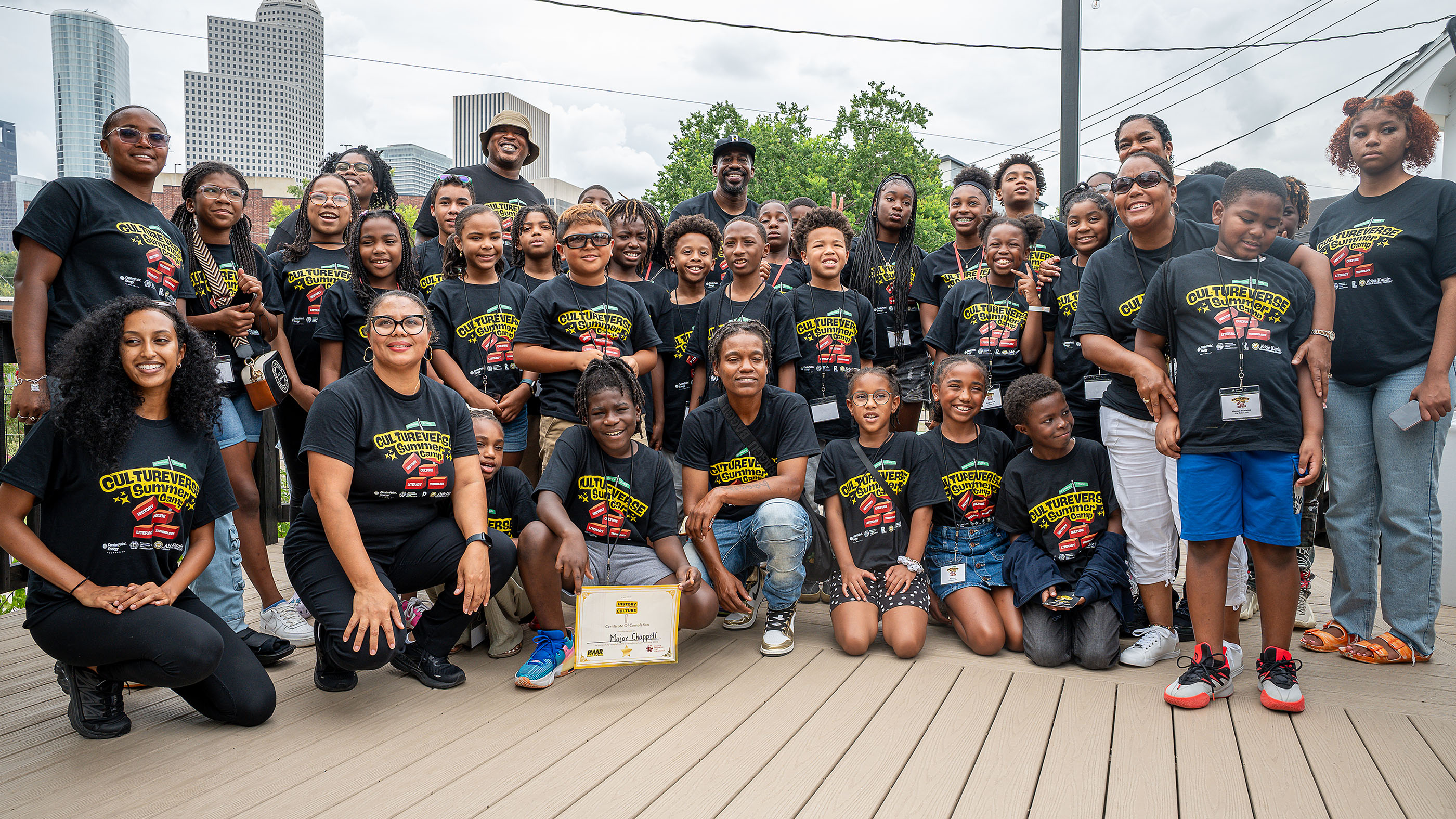 A diverse group of children and mentors in matching black Cultureverse T-shirts pose smiling on a wooden deck, a certificate visible.