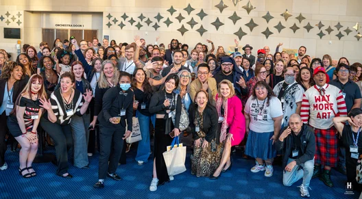 Large diverse crowd with name badges gathers in a lobby at Orchestra Door 1, many smiling and one person in red 'WAY MORE ART' sweater.