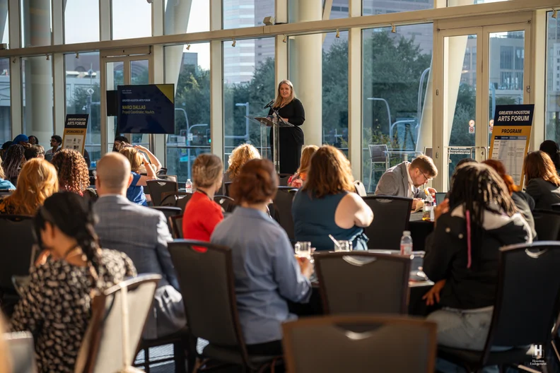 Sunlit glass-walled conference room with a female speaker at a clear podium addressing seated attendees at the Houston Arts Forum.