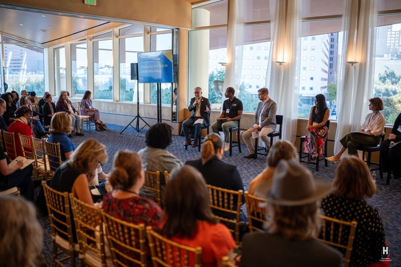 Panel discussion in a sunlit conference room with five seated panelists, an audience on gold chairs, large windows, and a screen.