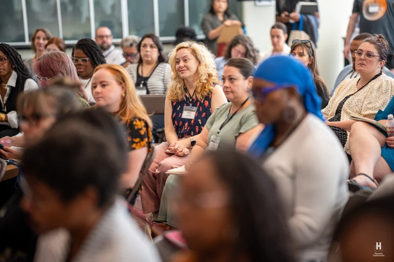 Diverse audience seated in a conference room, centered on a smiling blonde wearing a name badge, listening while others take notes.