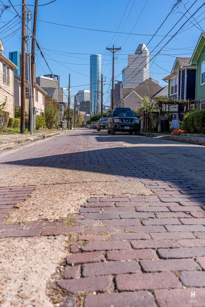 Low-angle view down a worn brick street with patchy asphalt, toward downtown skyscrapers, flanked by houses, utility poles and parked cars.