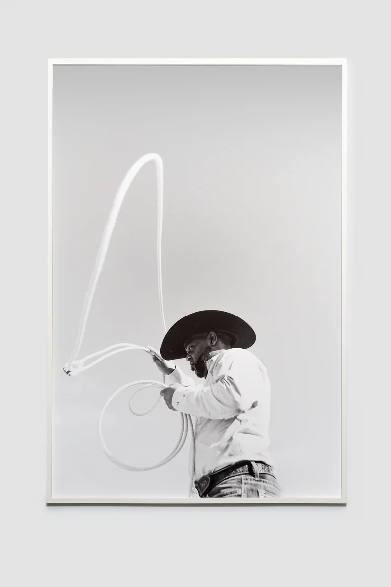 Framed black-and-white photo of a cowboy in a wide-brim hat, focused mid-throw as a lasso arcs overhead and coils at his waist.