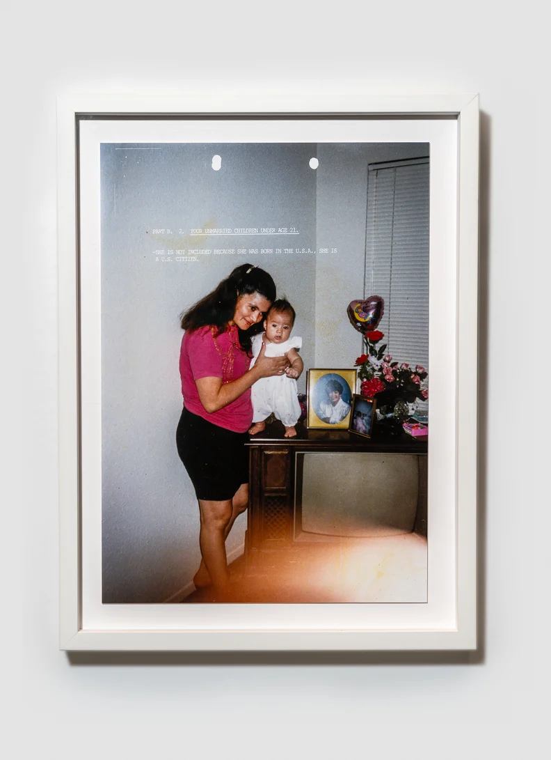 White-framed photograph shows a woman in pink holding a baby on a TV console beside framed photos, flowers and a heart-shaped balloon.