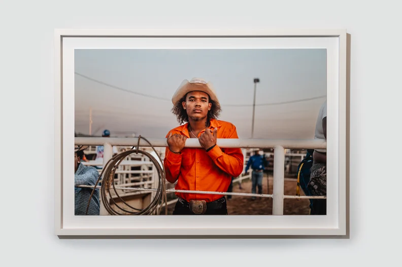 Young cowboy in a white hat and orange shirt grips metal fence at a rodeo pen, coiled lariat beside him at sunset, belt buckle visible.