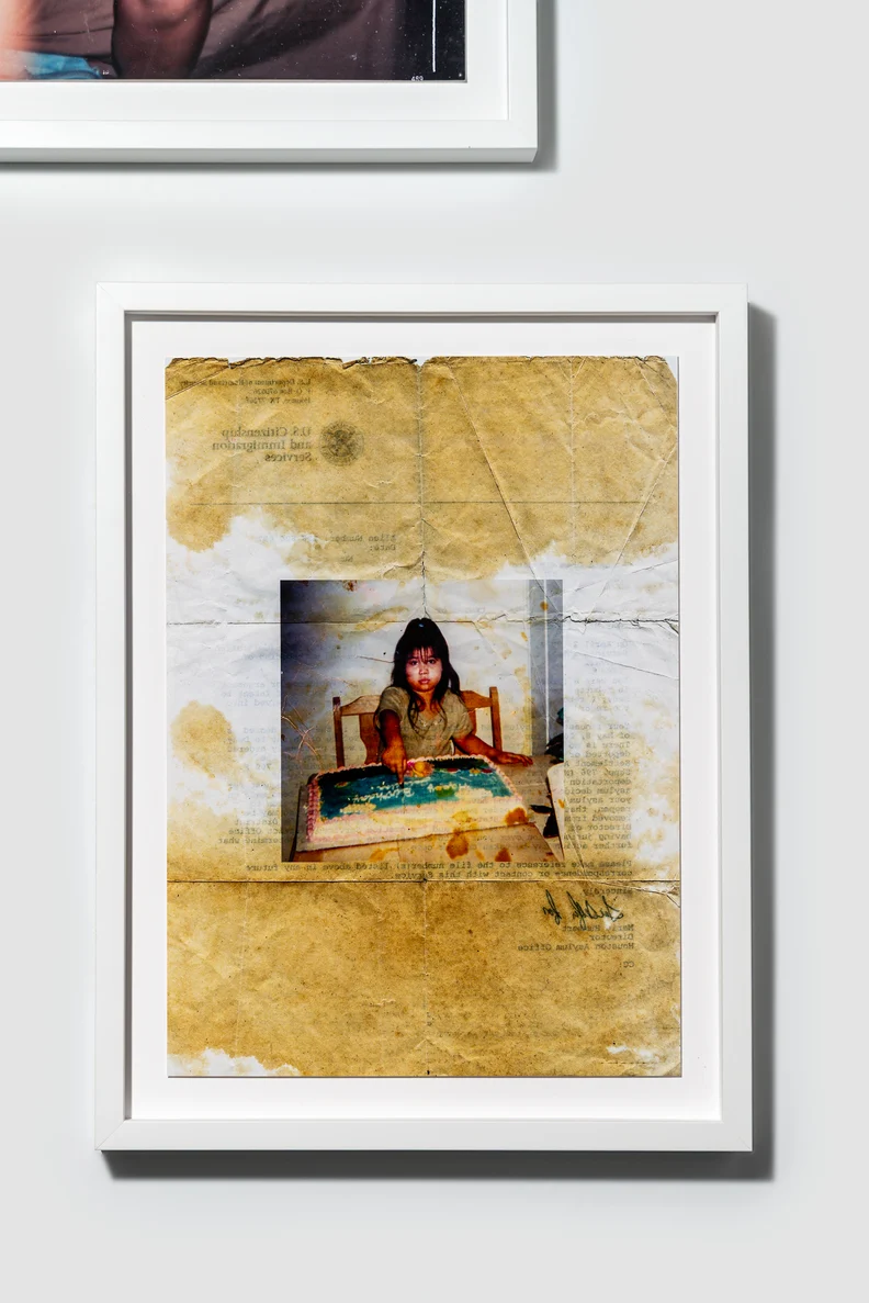 Framed, creased yellowed paper with stains around a small central photo of a young girl at a table pointing at a frosted birthday cake.