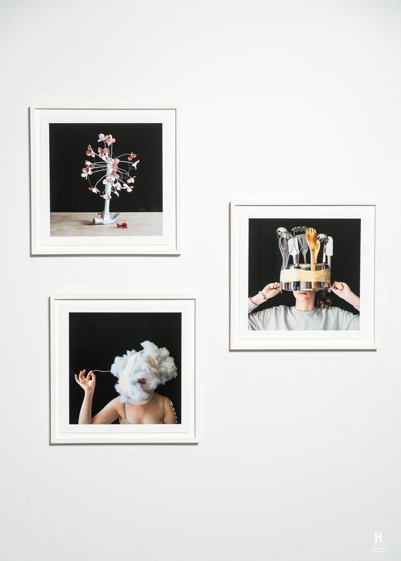 Three framed photos on white wall show a flower sculpture, a person wearing a pot of utensils on head, and a woman with a cotton cloud head.