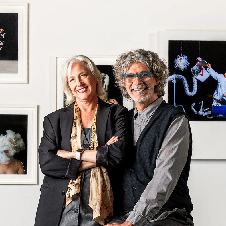 Smiling older woman with white hair in a black blazer and scarf poses with a gray-haired bespectacled man in a vest before framed art.