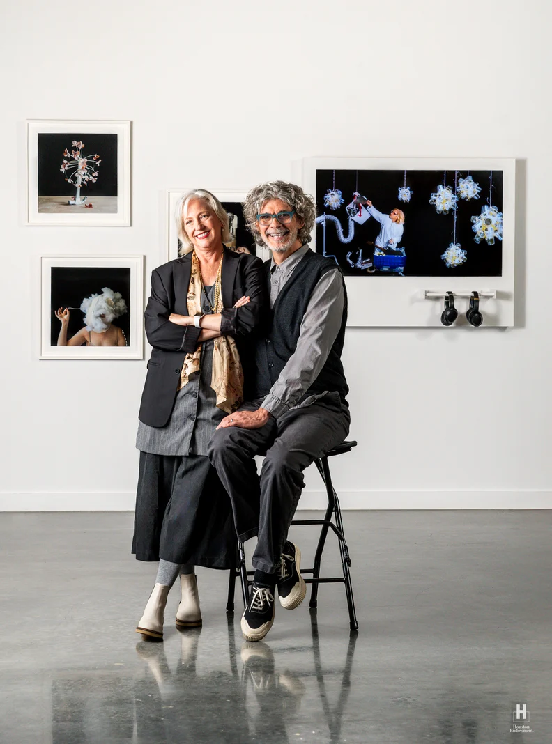 A smiling older woman stands with arms crossed next to a seated older man on a stool in a white gallery with framed photo art behind them.