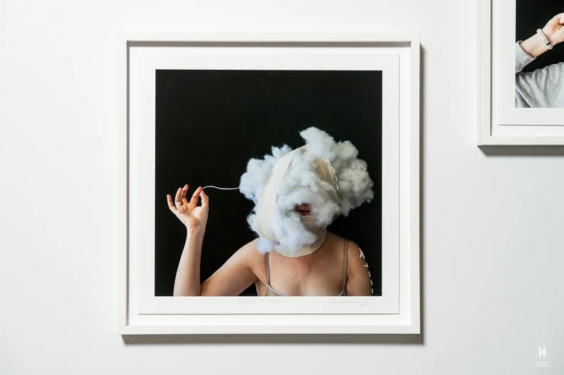 Framed photograph of a bare-shouldered person with bandages and a cotton cloud obscuring their face, tugging a thread.
