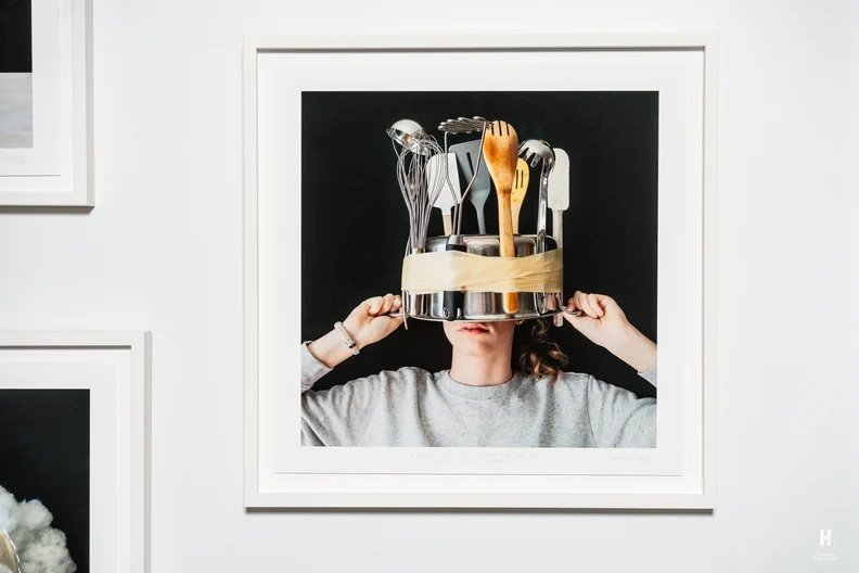Person in gray sweatshirt holds a metal pot taped over their head filled with wooden spoon, whisk, and spatulas, lower face visible.
