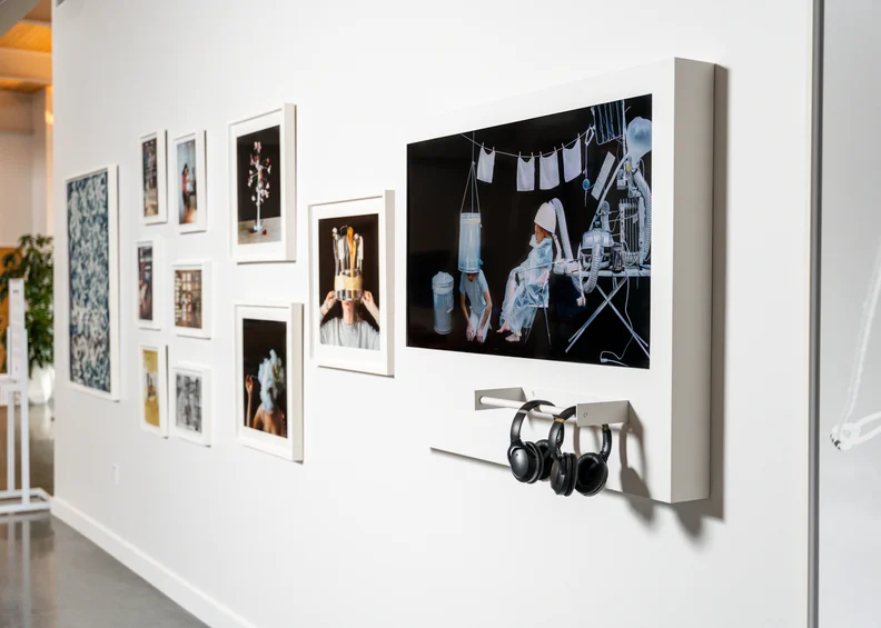 Gallery wall with framed photos and a mounted screen showing two figures in translucent gear under hung laundry, headphones hanging below.