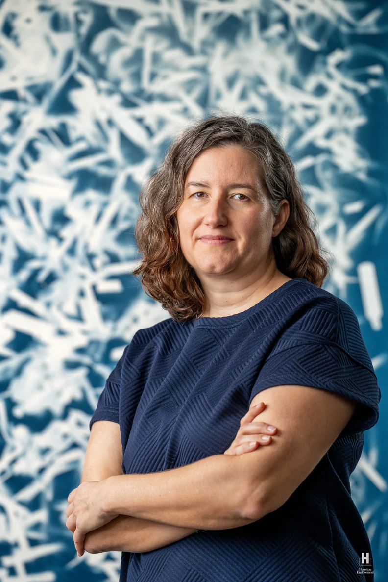 Middle-aged woman with shoulder-length wavy brown hair, arms crossed, wearing a navy shirt against a blue abstract background.