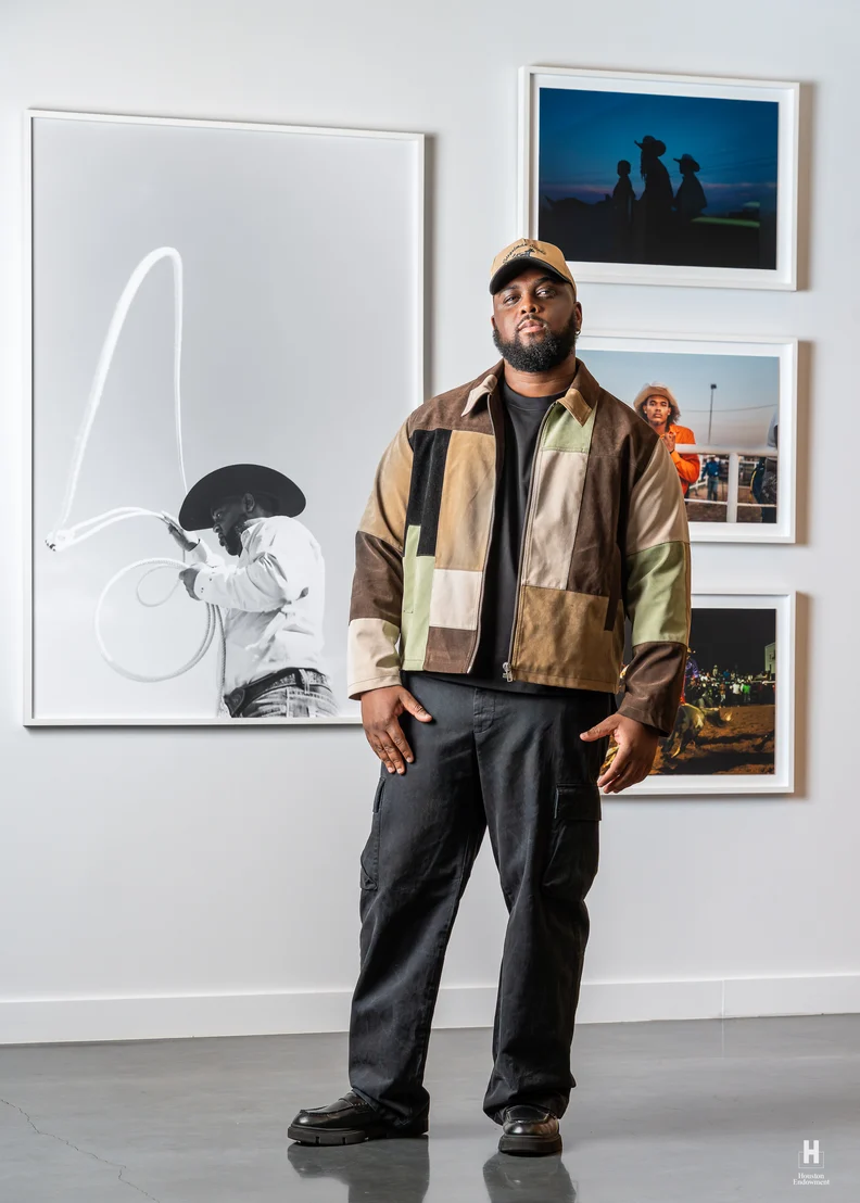 Bearded man in a patchwork jacket and cap stands in a gallery, hands at sides, before a large lasso photo and three framed color images.