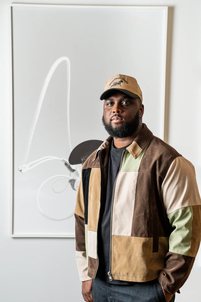 Bearded man wearing a beige cap and patchwork suede jacket stands relaxed before a large framed abstract monochrome artwork on wall.