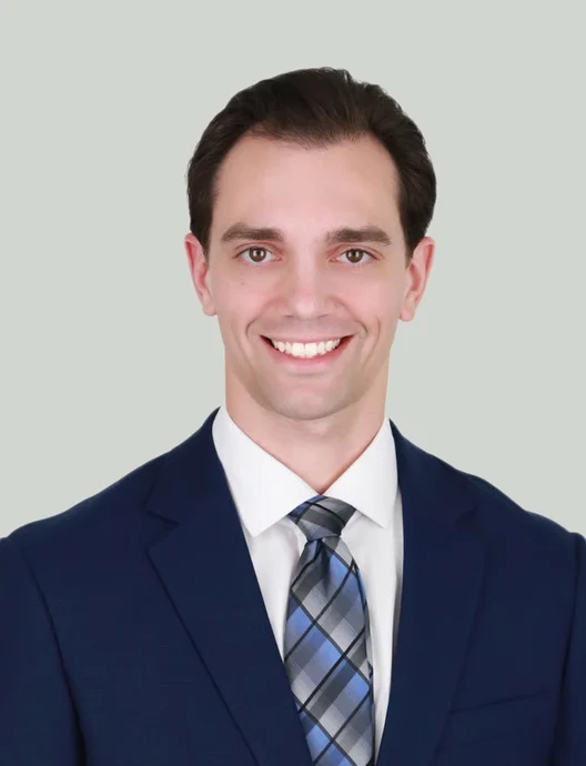 Smiling clean shaven man with dark hair in a navy suit, white shirt and blue-gray plaid tie, gazing at the camera against pale gray.